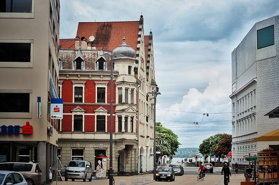 Rathausstraße in Bregenz: Bürobau, historistisches Eckhaus, Nachkriegsbau und das Vorarlberg Museum nebeneinander, dahinter der Bodensee